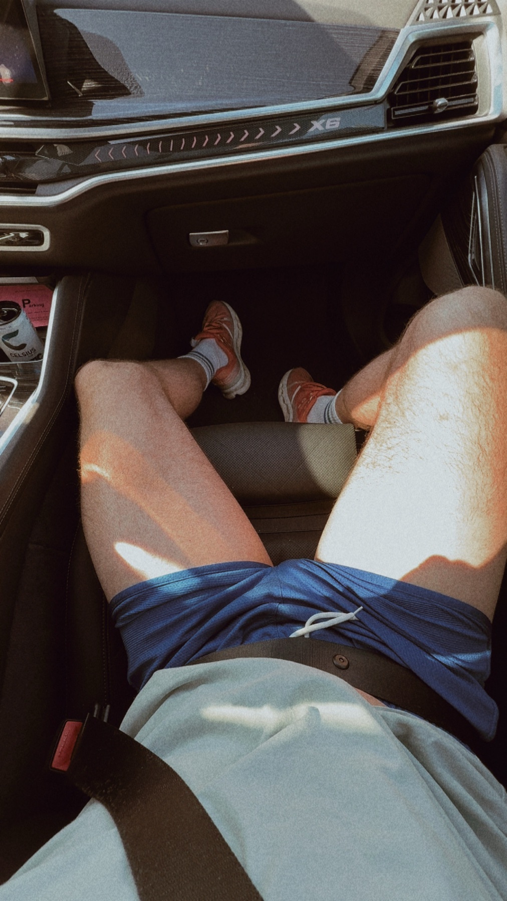 A man's legs are visible from the knees down, wearing blue shorts and sneakers, as he sits in the driver's seat of a car.