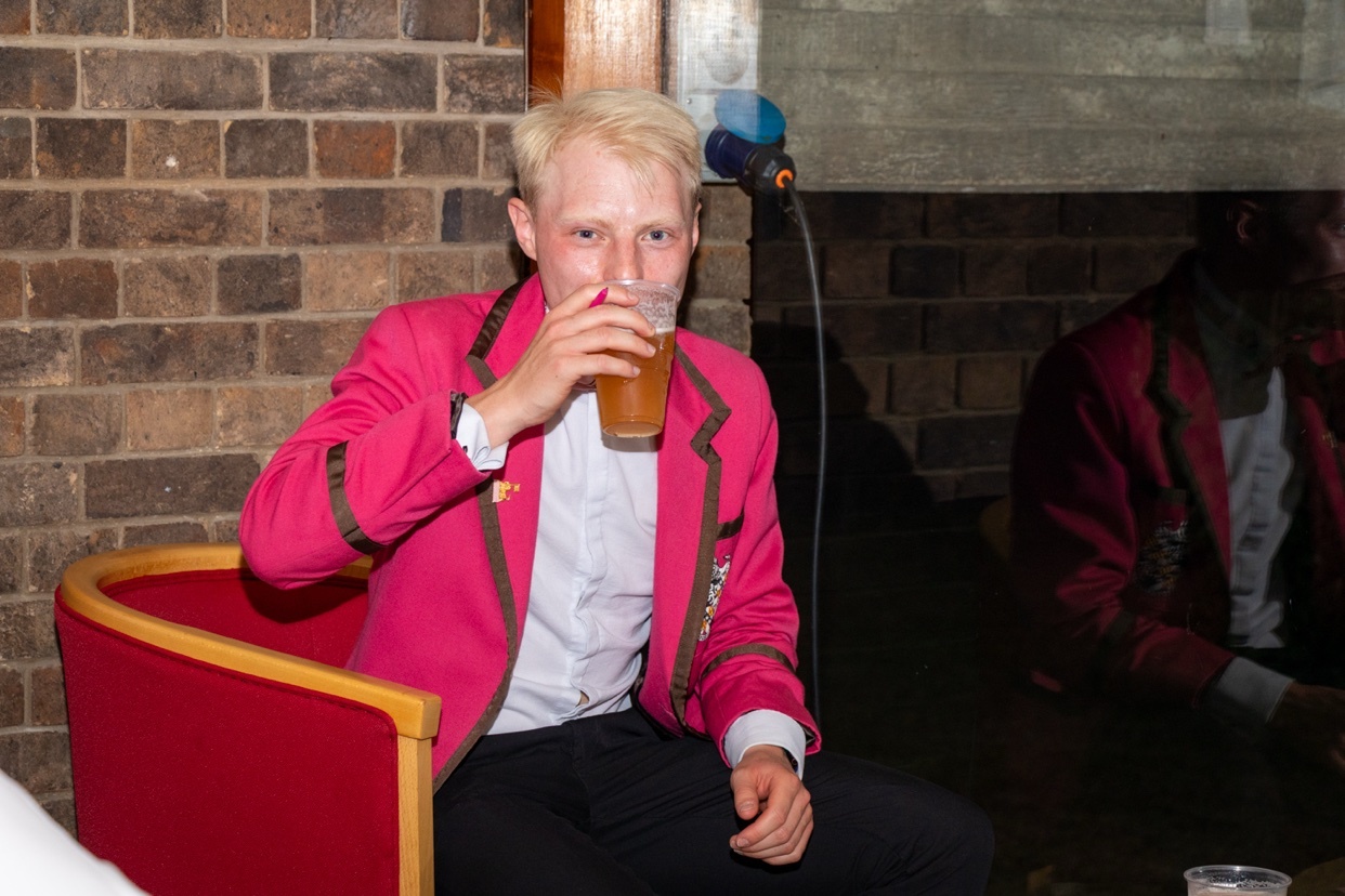 A man with blond hair is sitting indoors and drinking from a glass of beer. He is wearing a bright pink blazer over a white shirt and black pants.