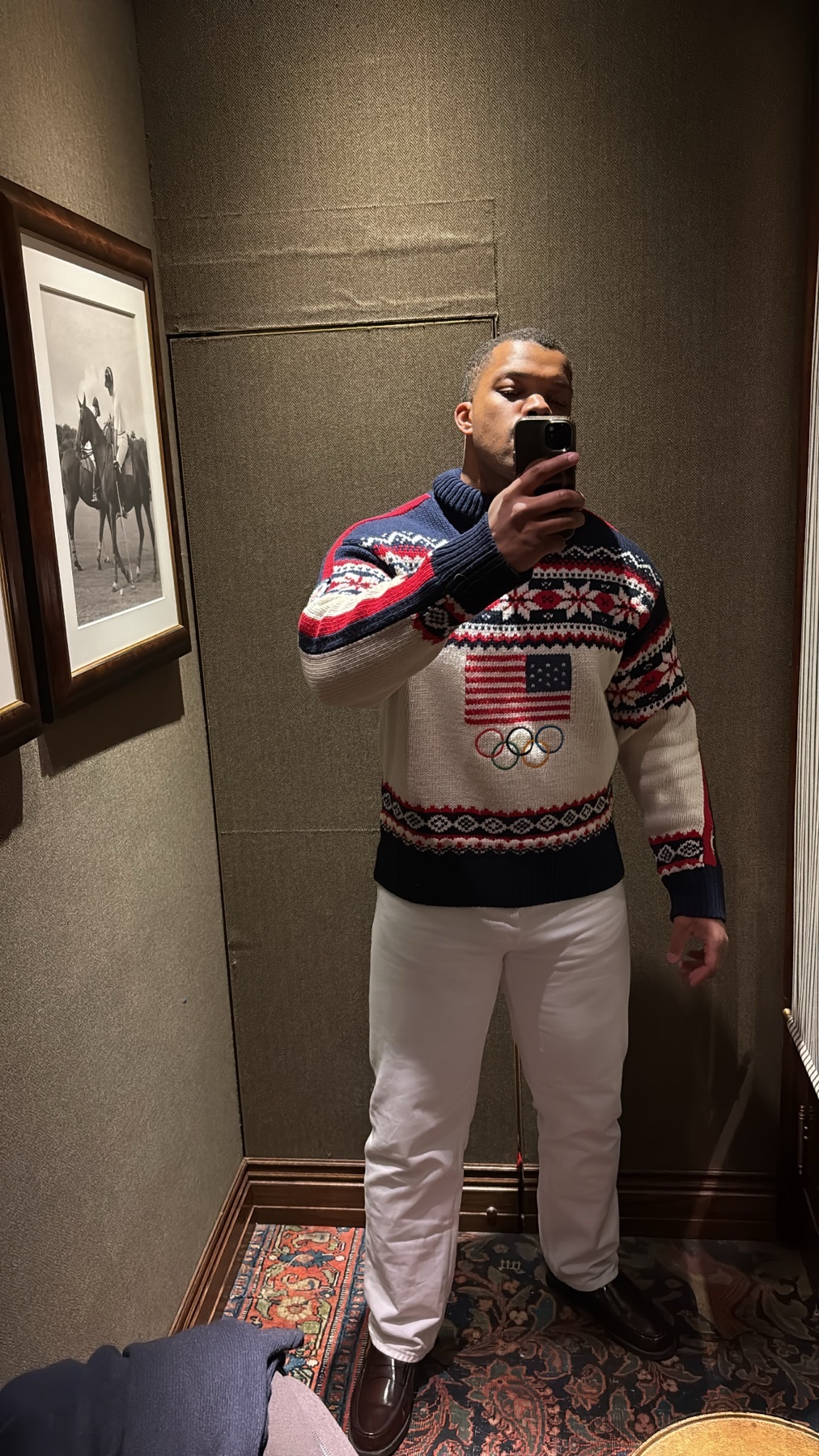 A man takes a selfie in a mirror, wearing a festive Ralph Lauren sweater adorned with the American flag and Olympic rings. He is also wearing white pants and brown dress shoes.