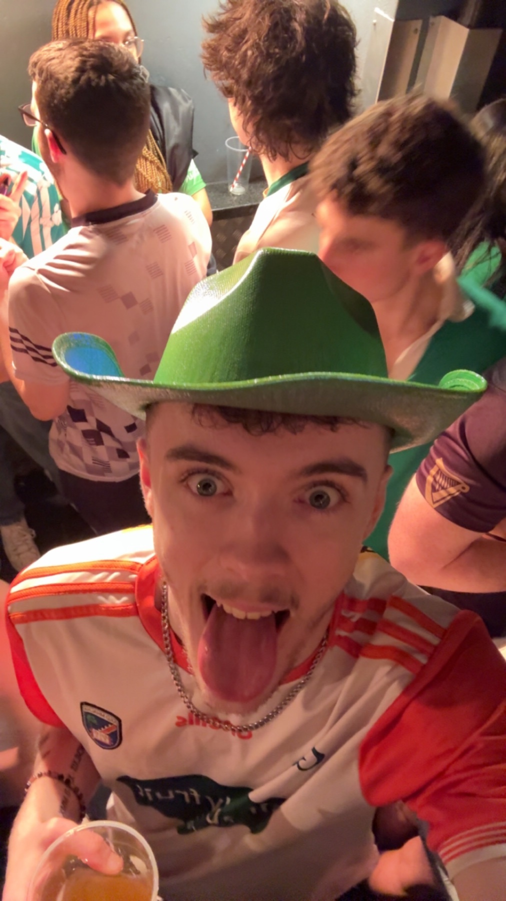 A man in a green cowboy hat sticks his tongue out at the camera while holding a drink at a lively party with friends.