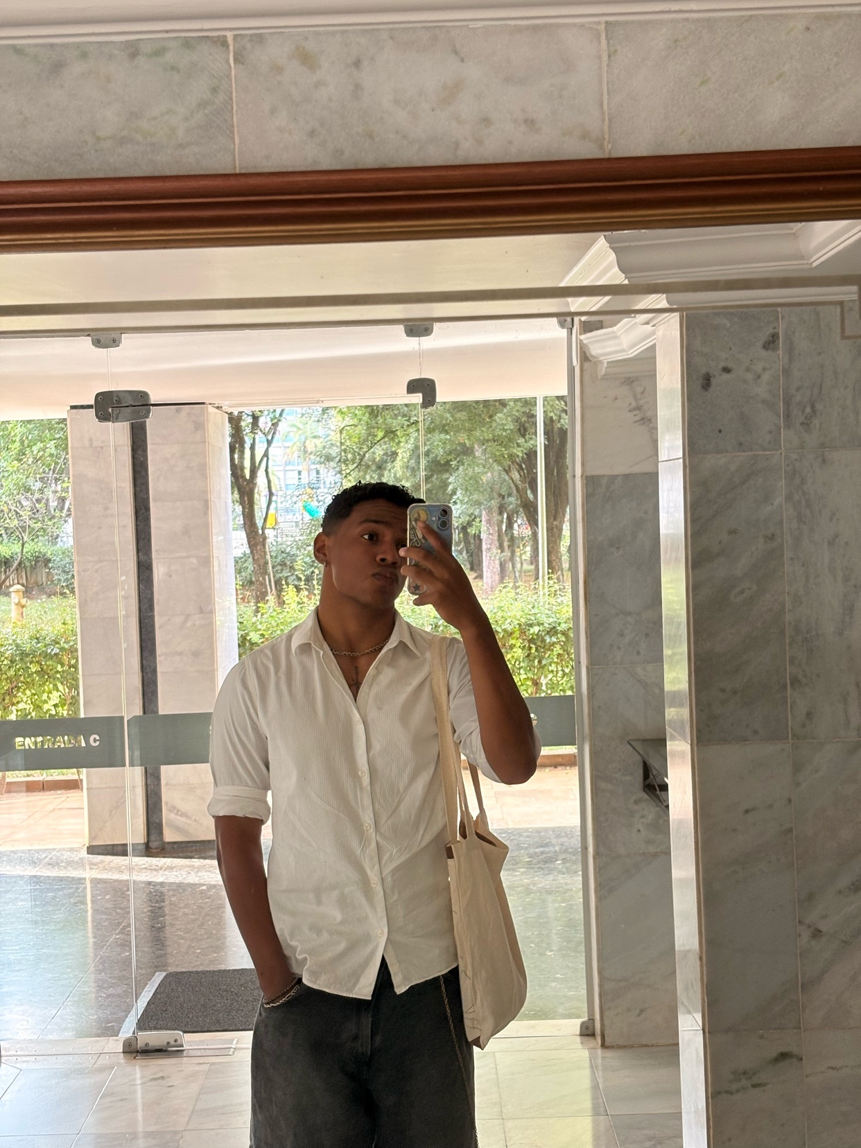 A man takes a mirror selfie in a white button-down shirt and dark pants, holding a canvas tote bag.