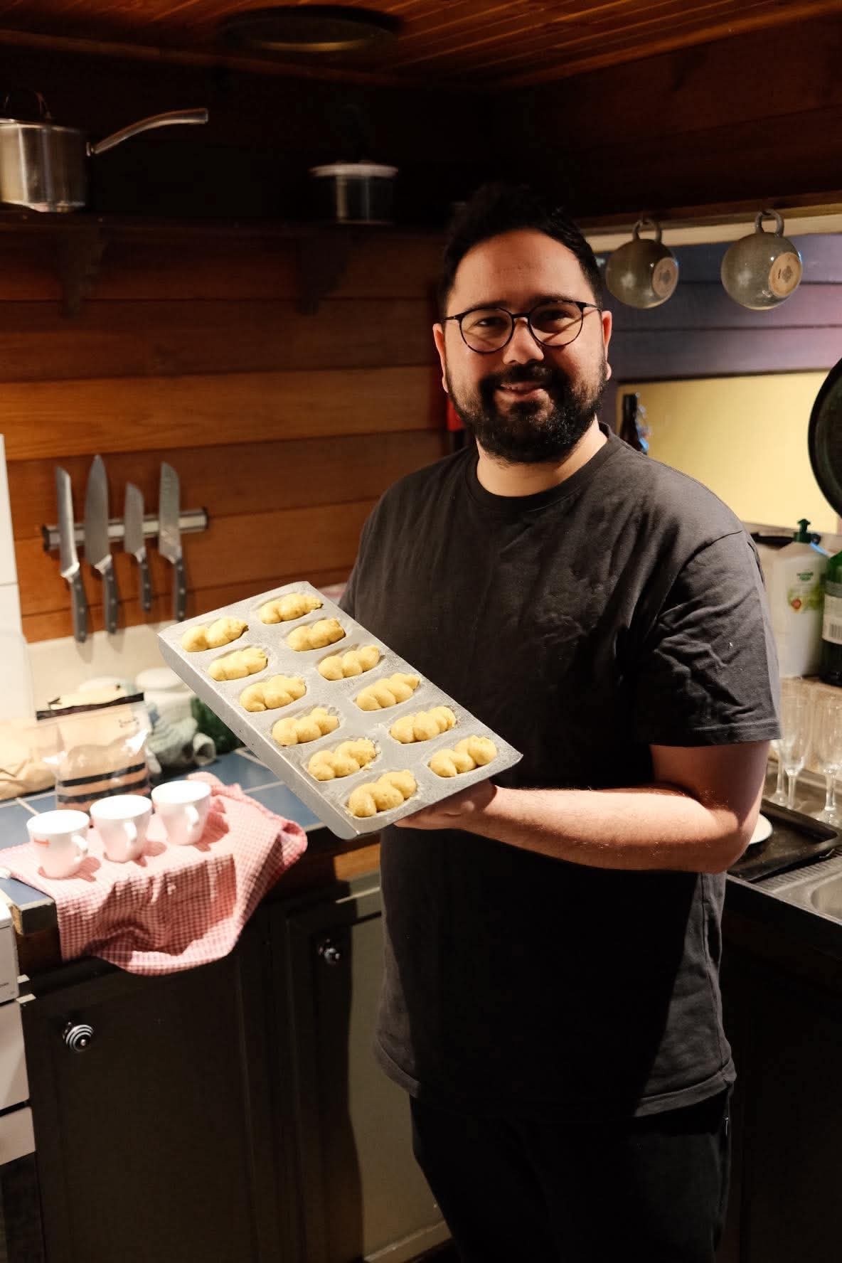 A man with glasses and a beard holds a baking tray filled with golden-brown madeleines. He is standing in a kitchen with wooden cabinets and a knife rack on the wall.