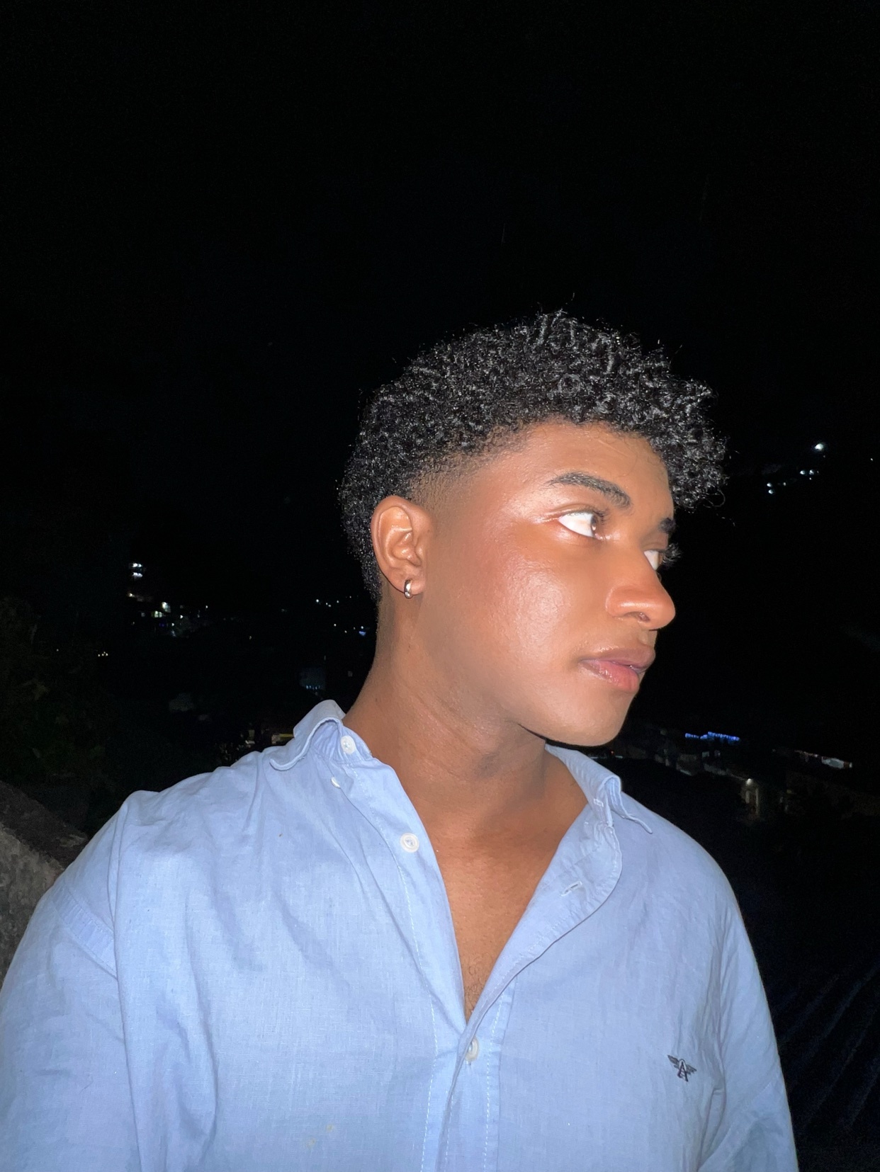 A man with curly hair poses for a photo at night. He is wearing a blue shirt and looking off to the side.