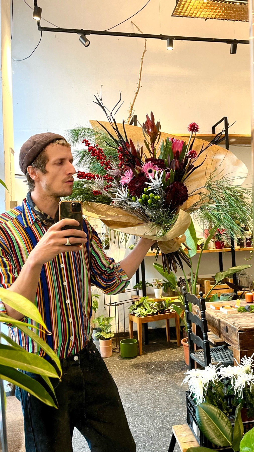 A man takes a mirror selfie, holding a large bouquet of flowers. He is wearing a colorful striped shirt and a cap.