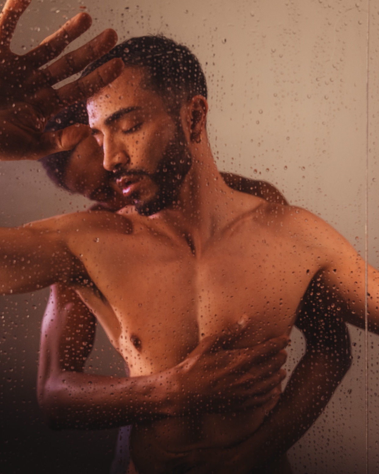 A shirtless man with his eyes closed is embraced by another person from behind, their hands resting on his chest. The image is taken through a glass surface covered in water droplets, creating a moody and intimate atmosphere.