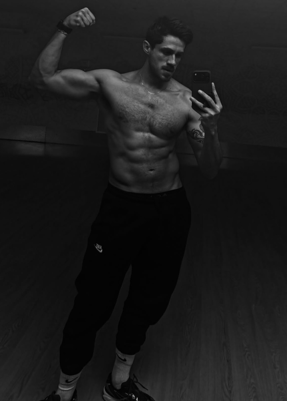 A man flexes his bicep in a mirror selfie, showcasing his muscular physique. The black and white photo highlights his defined abs and chest hair.