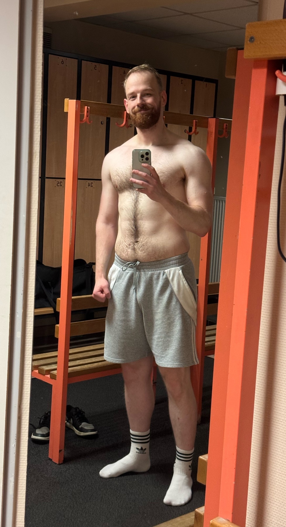A man takes a mirror selfie, shirtless, showing off his physique. He is wearing shorts and socks.