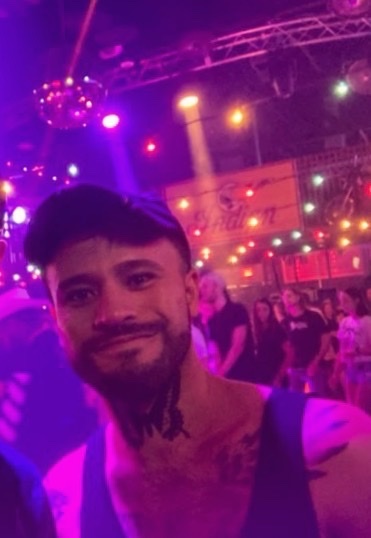 A man takes a selfie at a club, smiling at the camera. The background is blurred with a disco ball and other people.