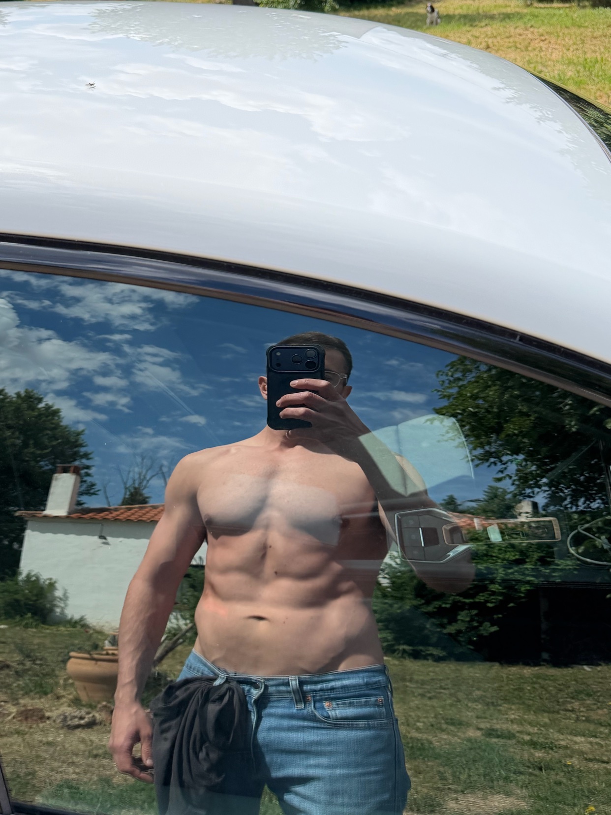A man with a muscular physique and visible abs takes a mirror selfie in a car. He is wearing jeans and has a towel tucked into his waistband.