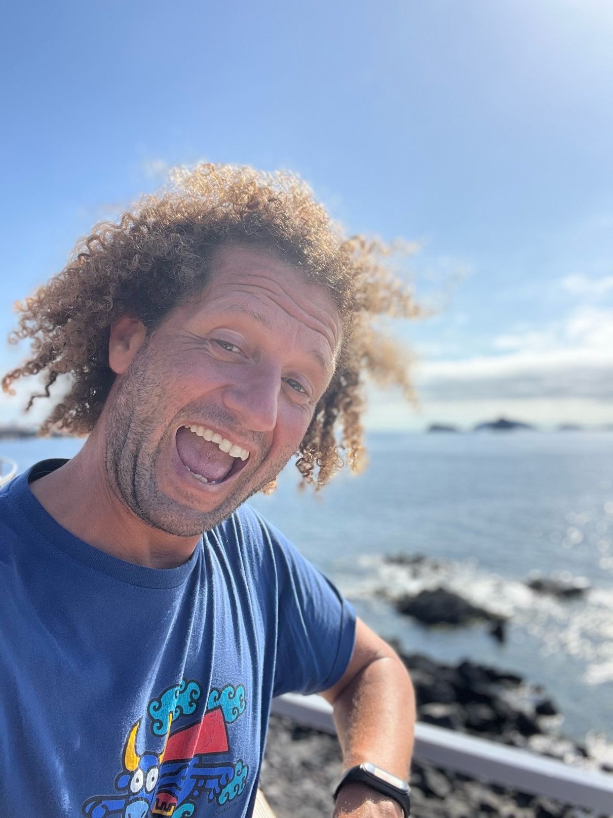 A man with curly hair smiles widely at the camera in a selfie taken outdoors with a blue t-shirt and the ocean in the background.