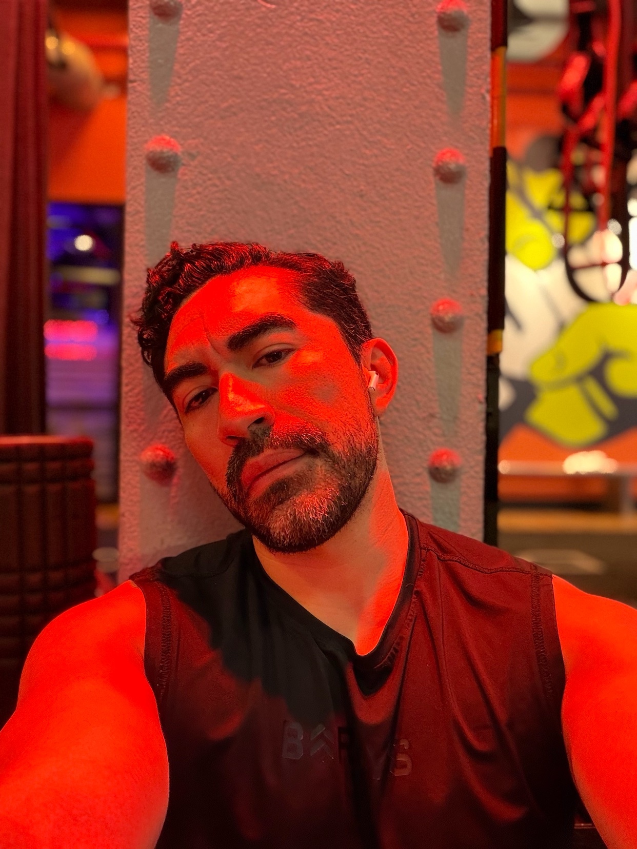 A man with a beard and earbuds poses for a selfie, leaning against a pillar under red lighting. Graffiti art is visible in the background.