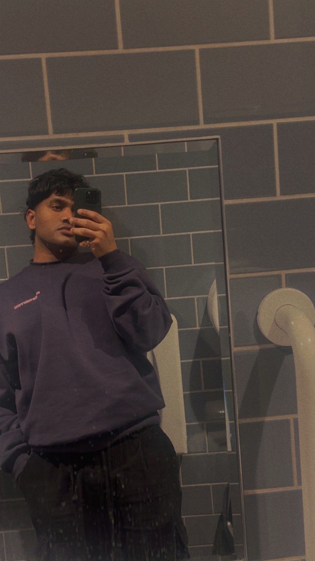 A man takes a mirror selfie in a bathroom. He is wearing a purple sweatshirt and black pants.