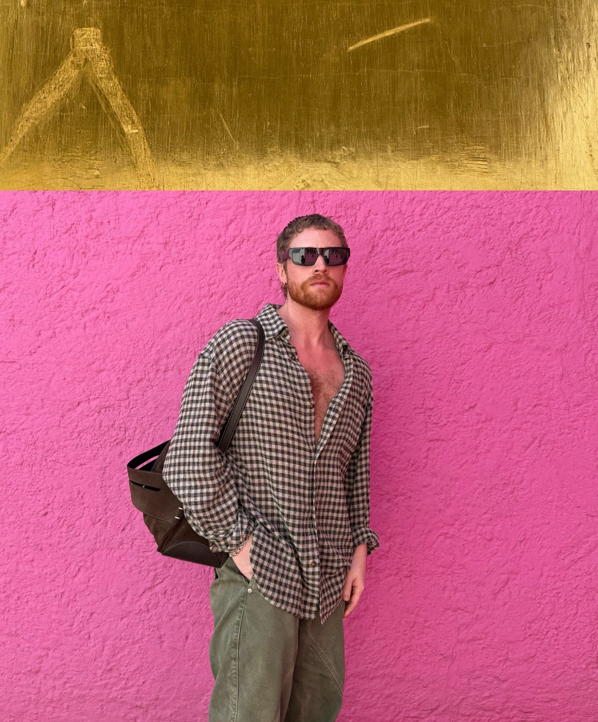 A man poses in front of a pink wall, wearing sunglasses and an unbuttoned shirt. His chest hair is visible.