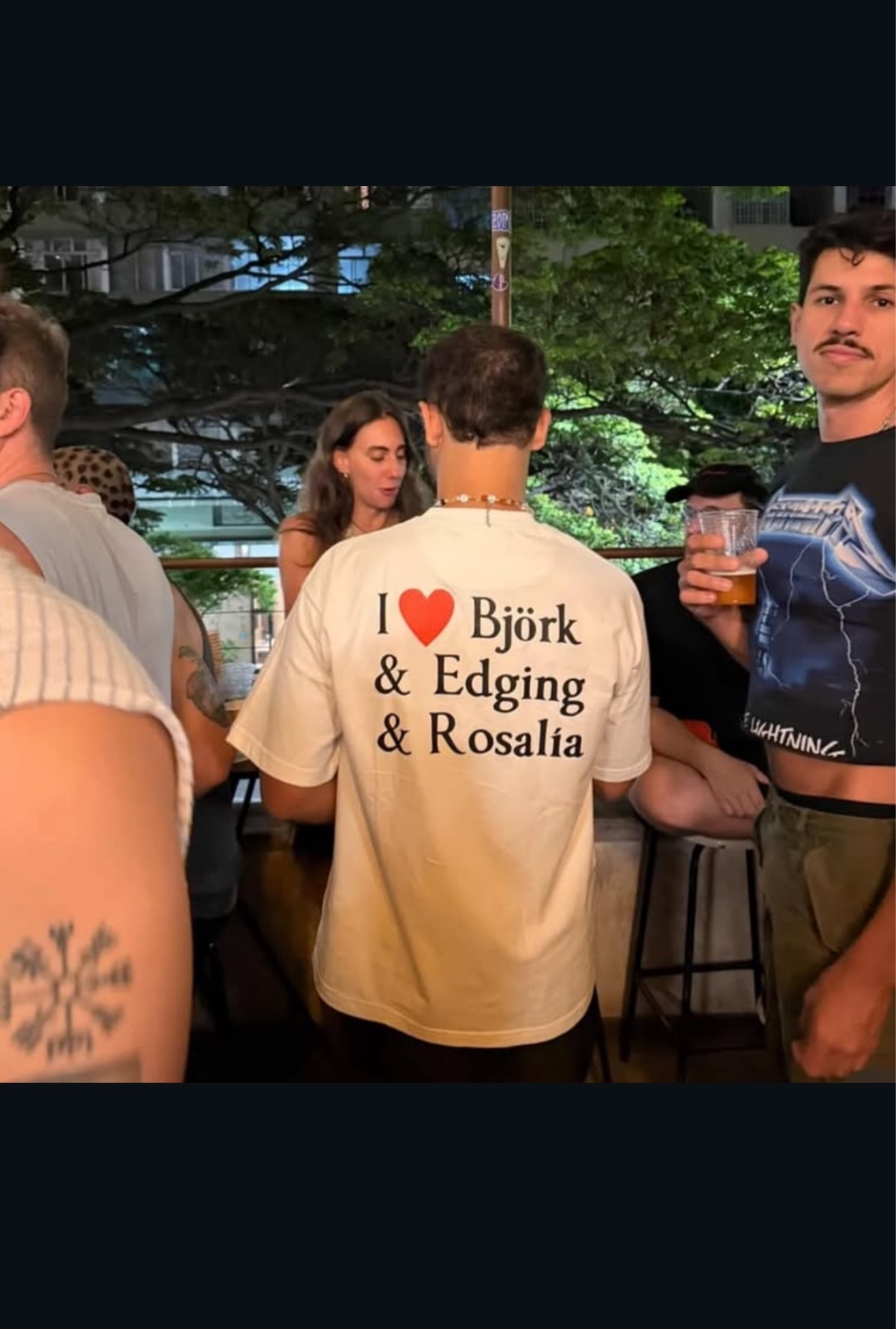 A man in a white t-shirt with the text "I ❤️ Björk & Edging & Rosalía" on the back is seen from behind. He is standing with a group of people, one of whom is holding a beer and has a mustache. The man is wearing a beaded necklace.