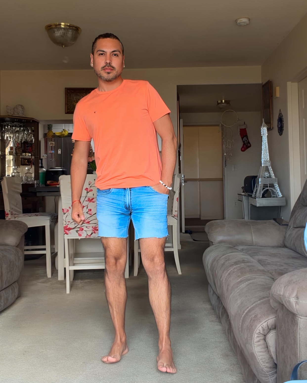A man stands in a living room wearing a bright orange t-shirt and light blue denim shorts. He is barefoot and has his left hand in his pocket.