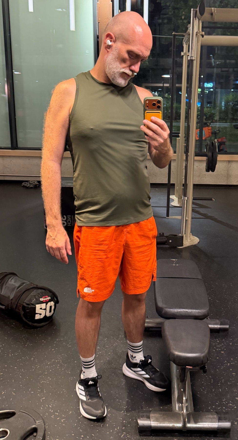 A bald man with a grey beard takes a mirror selfie in a gym. He is wearing an olive green tank top and bright orange athletic shorts.