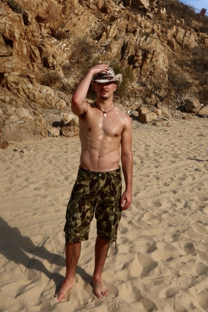 A shirtless man wearing a cowboy hat and camouflage shorts stands on a sandy beach, with a rocky hillside in the background.