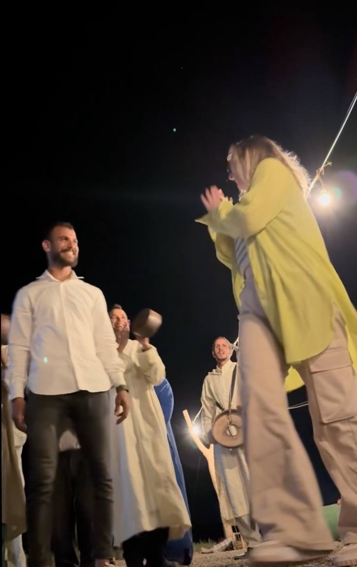 A group of people are gathered outdoors at night, with one woman in a bright yellow coat and another person in a white robe holding a small instrument. The man in the foreground wears a white button-down shirt and dark pants, smiling as he looks towa