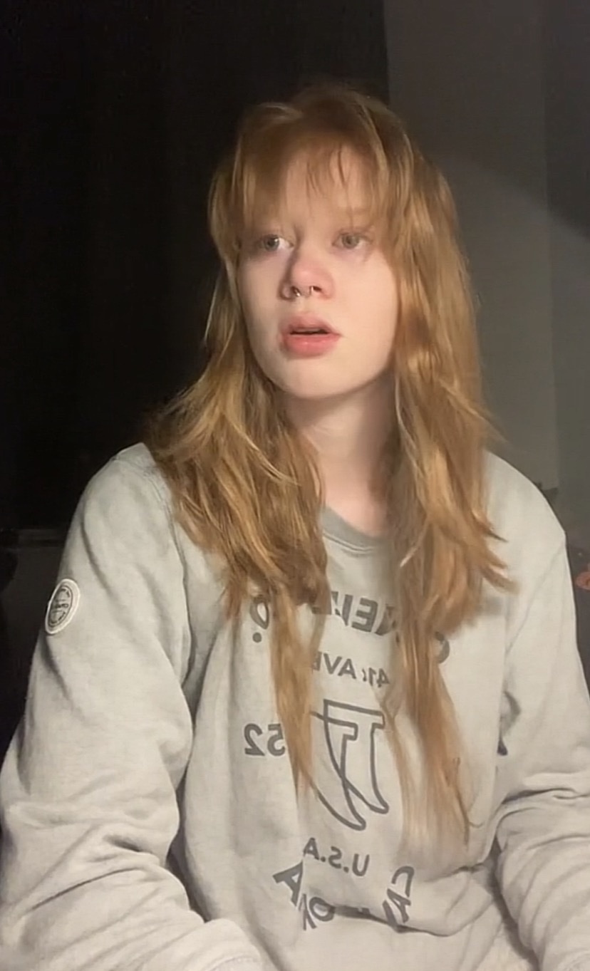 A young woman with long, blonde hair and bangs looks off to the side with a concerned expression. She is wearing a grey sweatshirt.