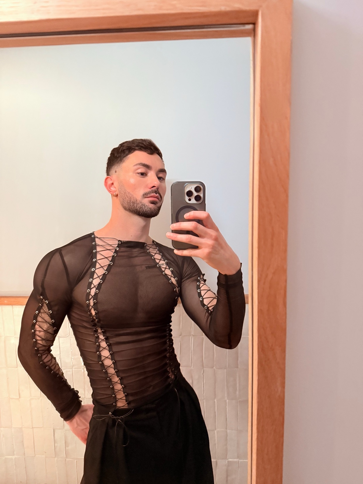 A man takes a mirror selfie, showcasing his physique and fashion sense. He is shirtless, wearing a stylish top and black pants.