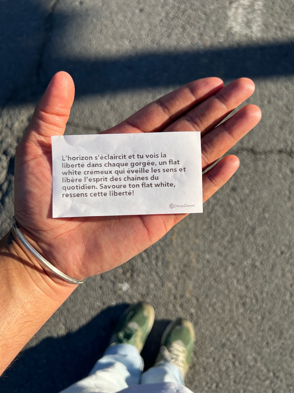 A hand holds a small piece of paper with a poem written in French about freedom and enjoying a flat white. The hand is wearing a bracelet, and the person's feet in sneakers are visible below.