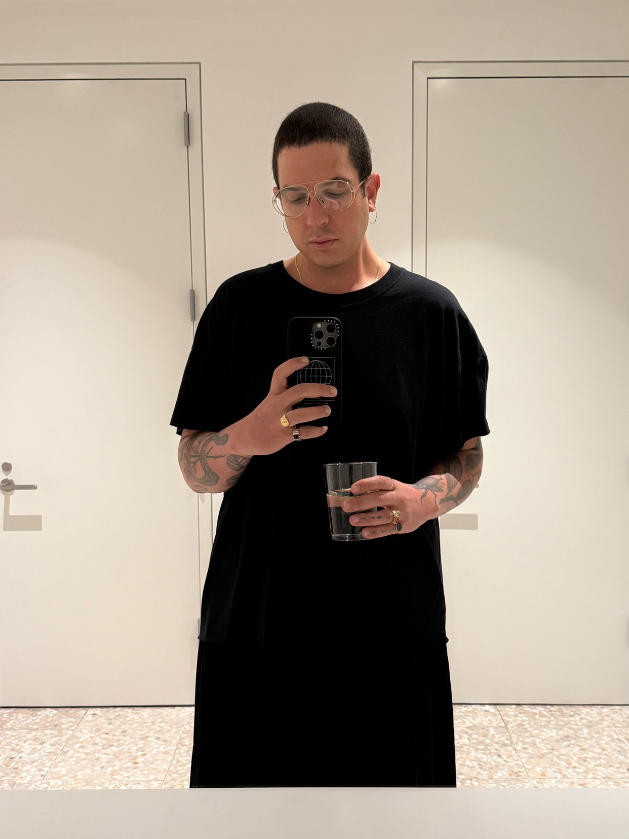 A man takes a mirror selfie, holding a phone and a glass. He wears glasses and a black t-shirt.