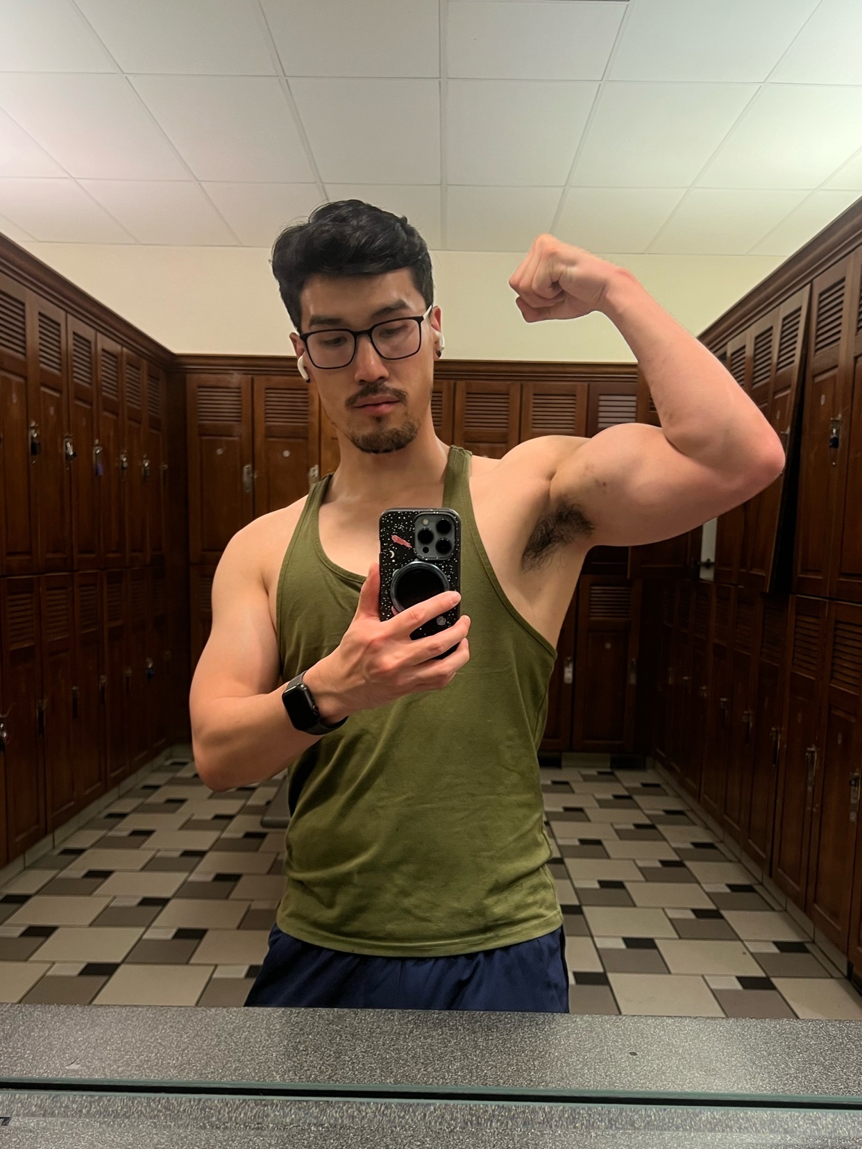 A man flexes his bicep in a gym mirror. He is wearing a tank top and glasses.