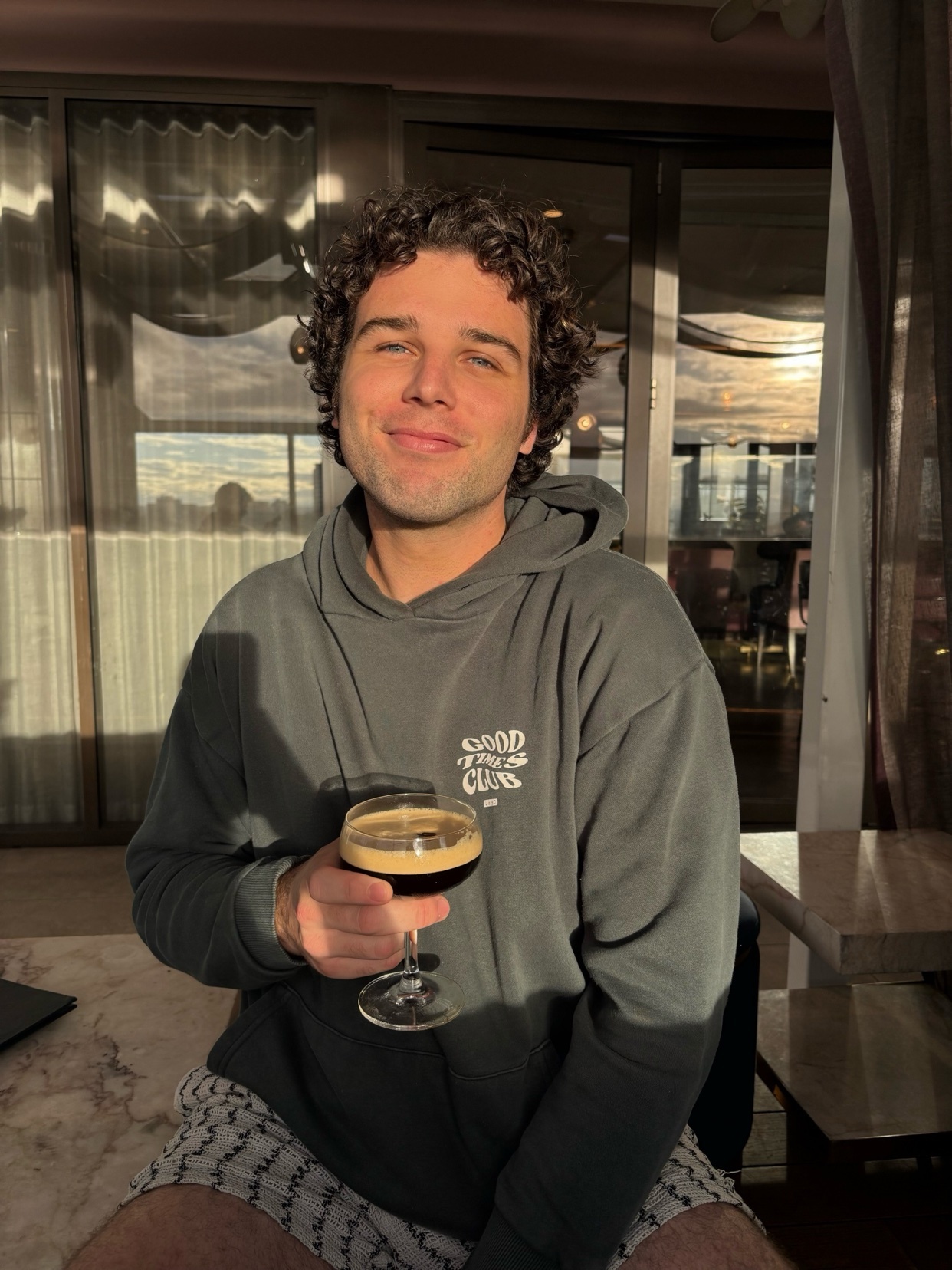 A man with curly hair smiles while holding a drink in a martini glass. He is wearing a dark hoodie and patterned shorts.
