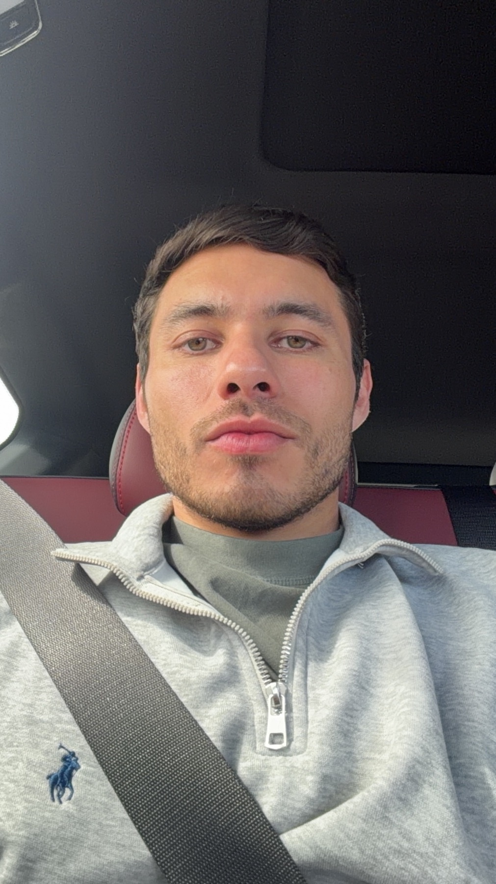 A man takes a selfie in the driver's seat of a car, wearing a grey quarter-zip polo shirt over a green t-shirt. He has stubble and is looking directly at the camera.