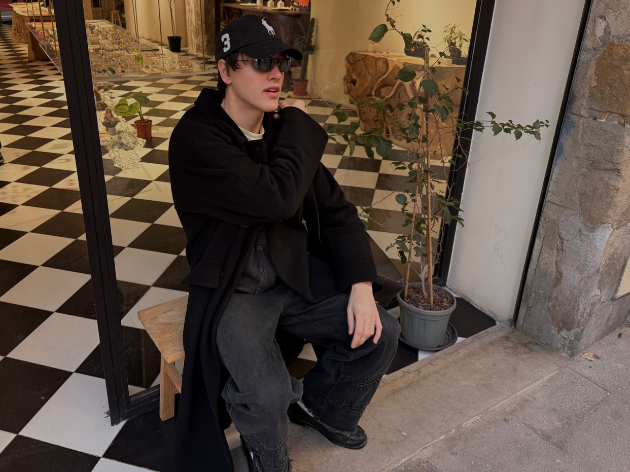 A man poses in a stylish black coat and cap. The photo is taken in front of a building with a checkered floor.