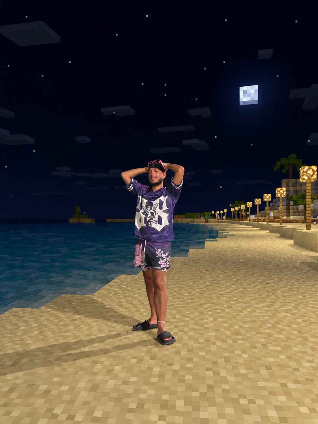 A man stands on a pixelated beach at night, with a full moon in the sky. The scene is reminiscent of the video game Minecraft, with blocky textures and a starry night sky.
