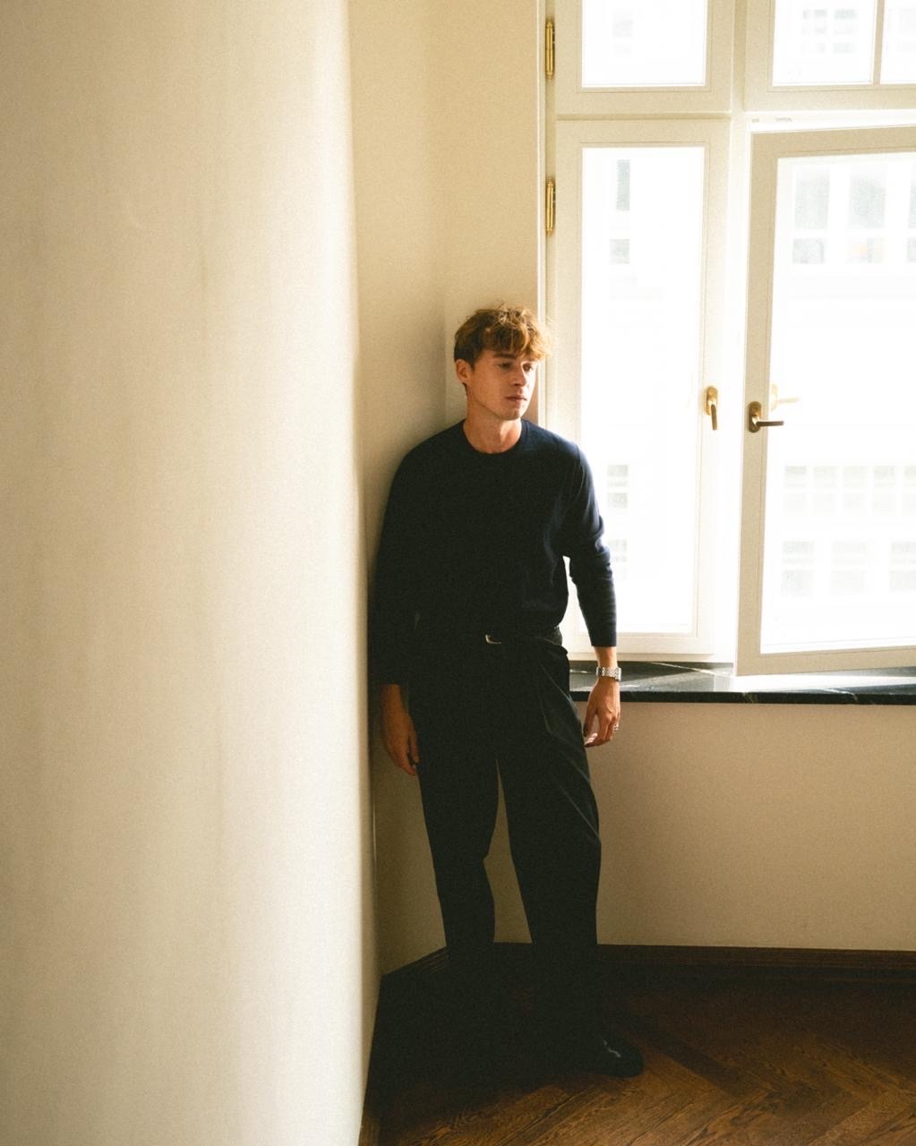 A man with curly hair stands indoors near a window, looking away from the camera. He is dressed in a dark sweater and dark pants.