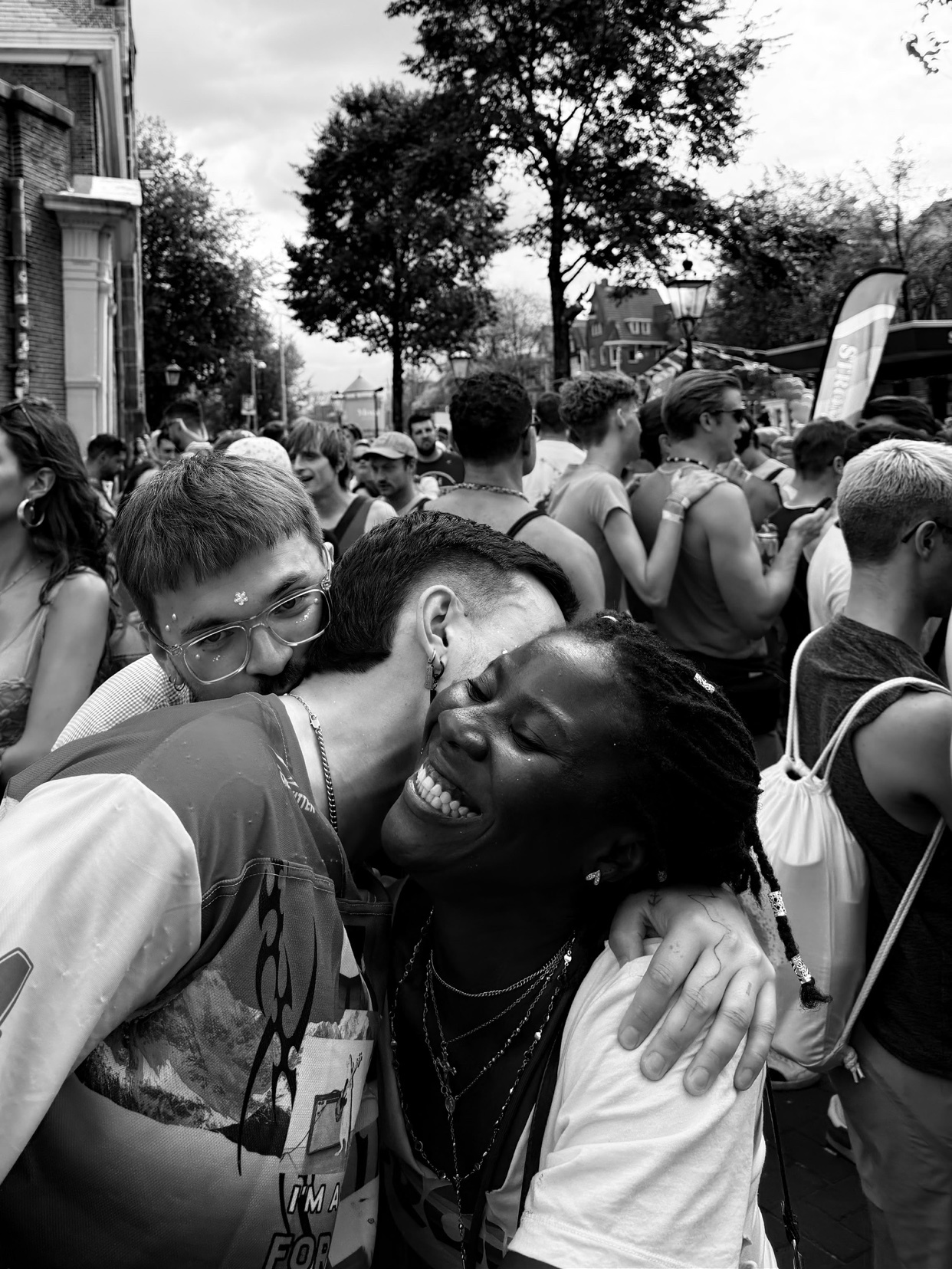 A black woman laughs joyfully as a man whispers in her ear in this black and white photo. They are surrounded by a crowd of people at an outdoor event.