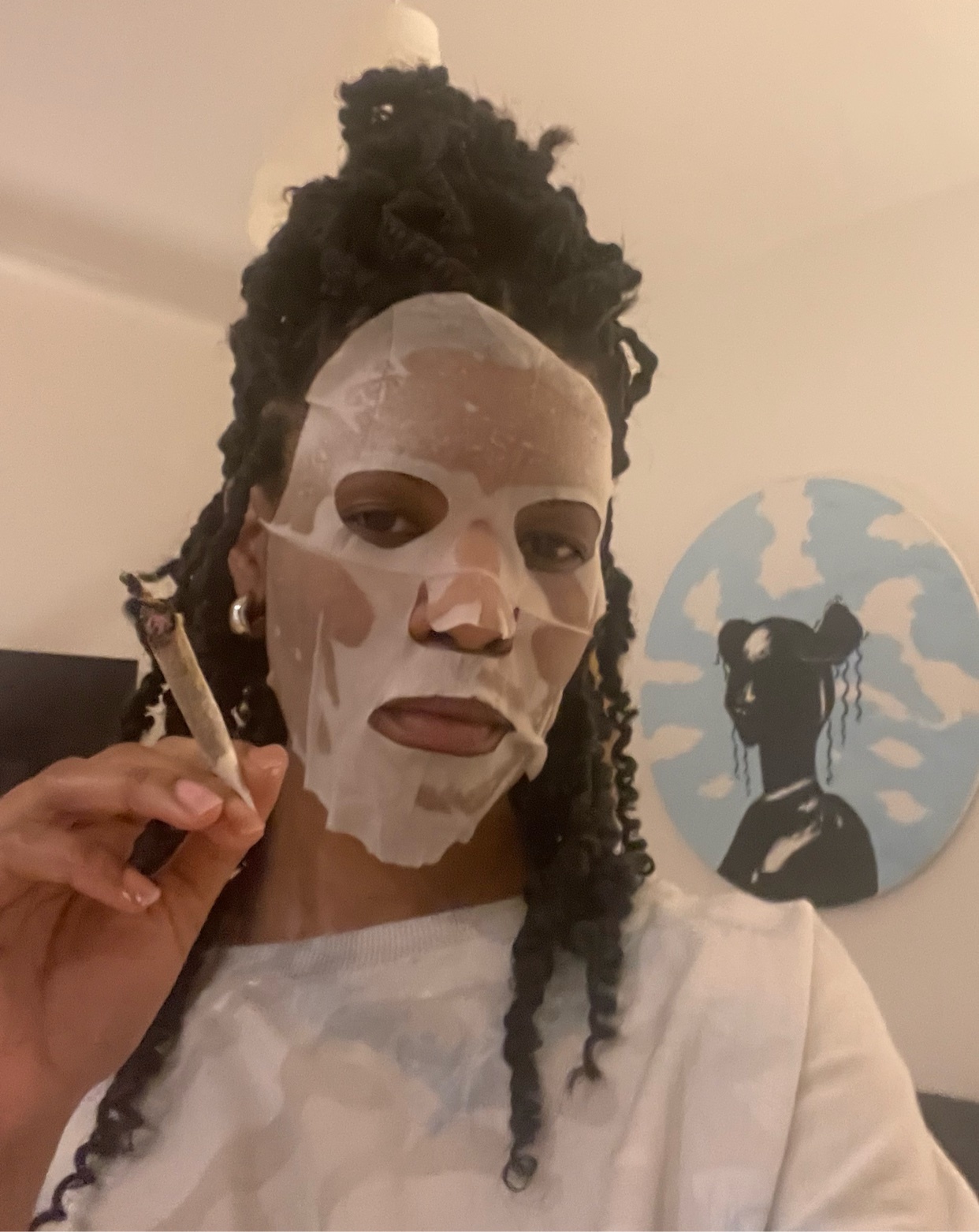 A woman with curly black hair and a face mask holds a lit joint. A piece of art is visible in the background.