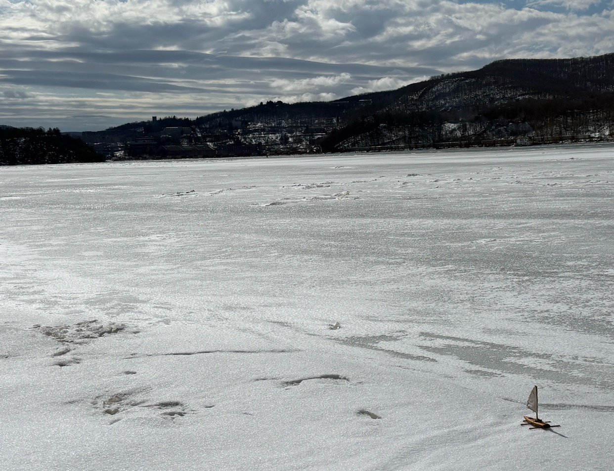 A small toy sailboat glides across a vast, frozen body of water under a cloudy sky. The icy landscape stretches towards a distant, tree-covered hill.