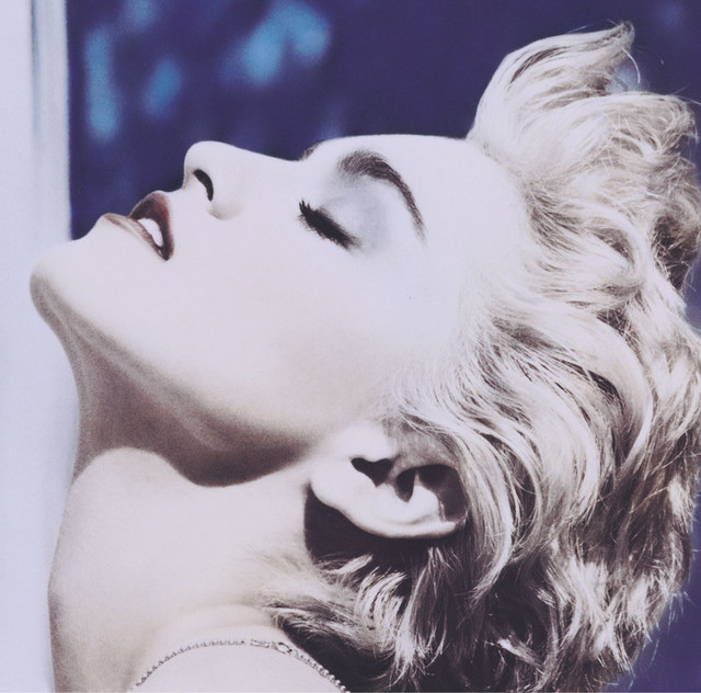 A close-up profile shot of a woman with blonde, curly, short hair, eyes closed, and head tilted back. Her makeup is dramatic, with dark lipstick and eyeshadow.