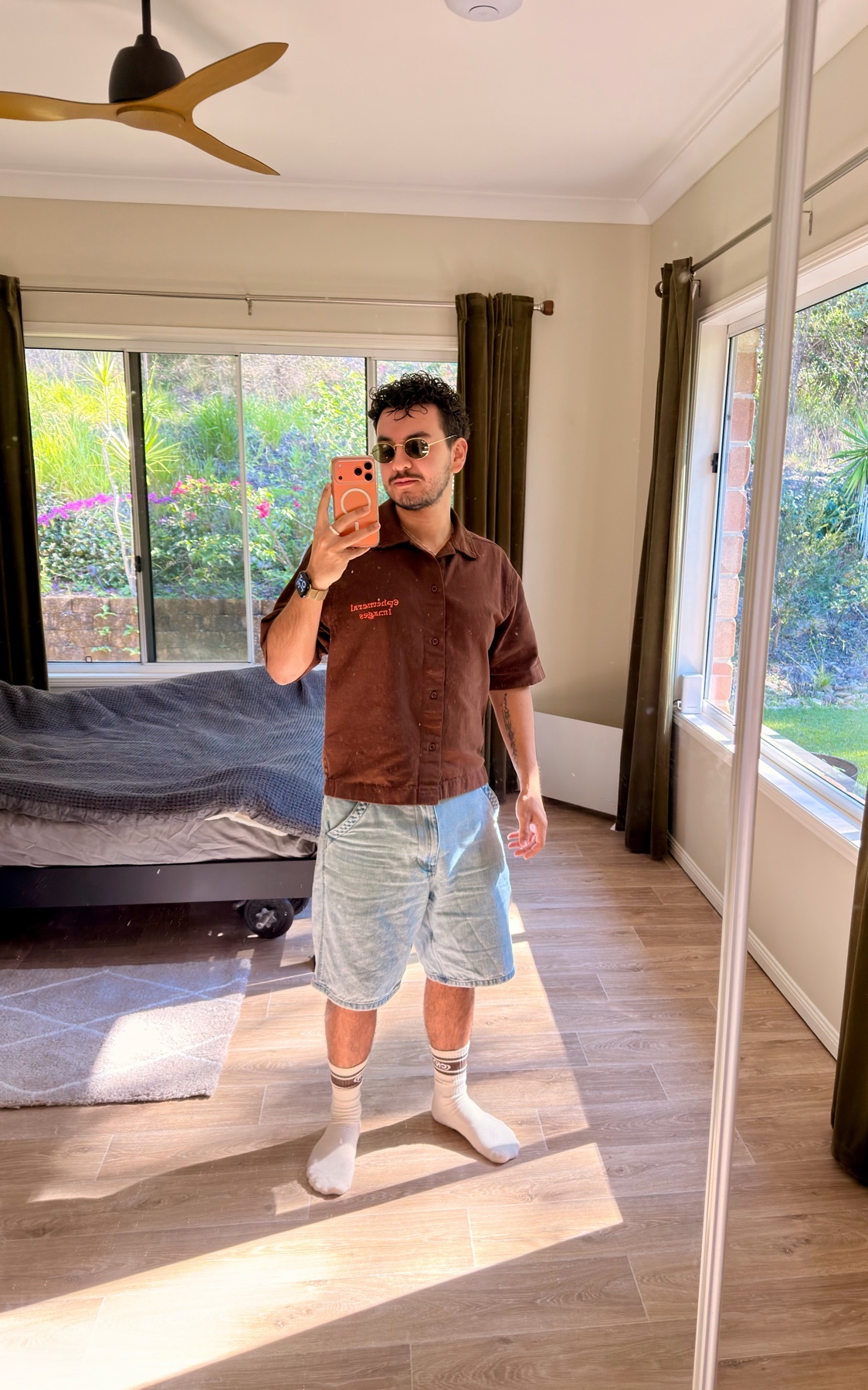 A man takes a mirror selfie in a bedroom, wearing sunglasses, a brown short-sleeved shirt, light-wash denim shorts, and white socks.