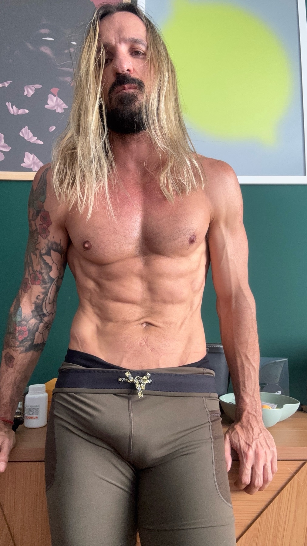 A man with long blonde hair and a muscular physique poses shirtless. He is wearing athletic shorts and has tattoos on his arm.