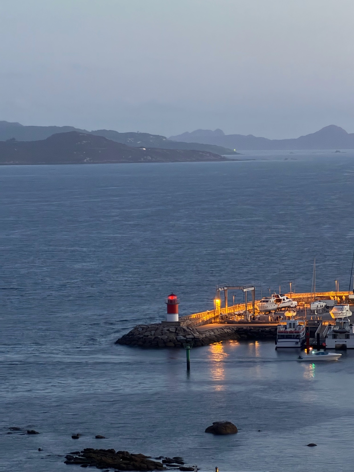 A lighthouse stands guard over a small harbor filled with boats at dusk. The warm lights of the harbor reflect on the calm water, creating a serene maritime scene.