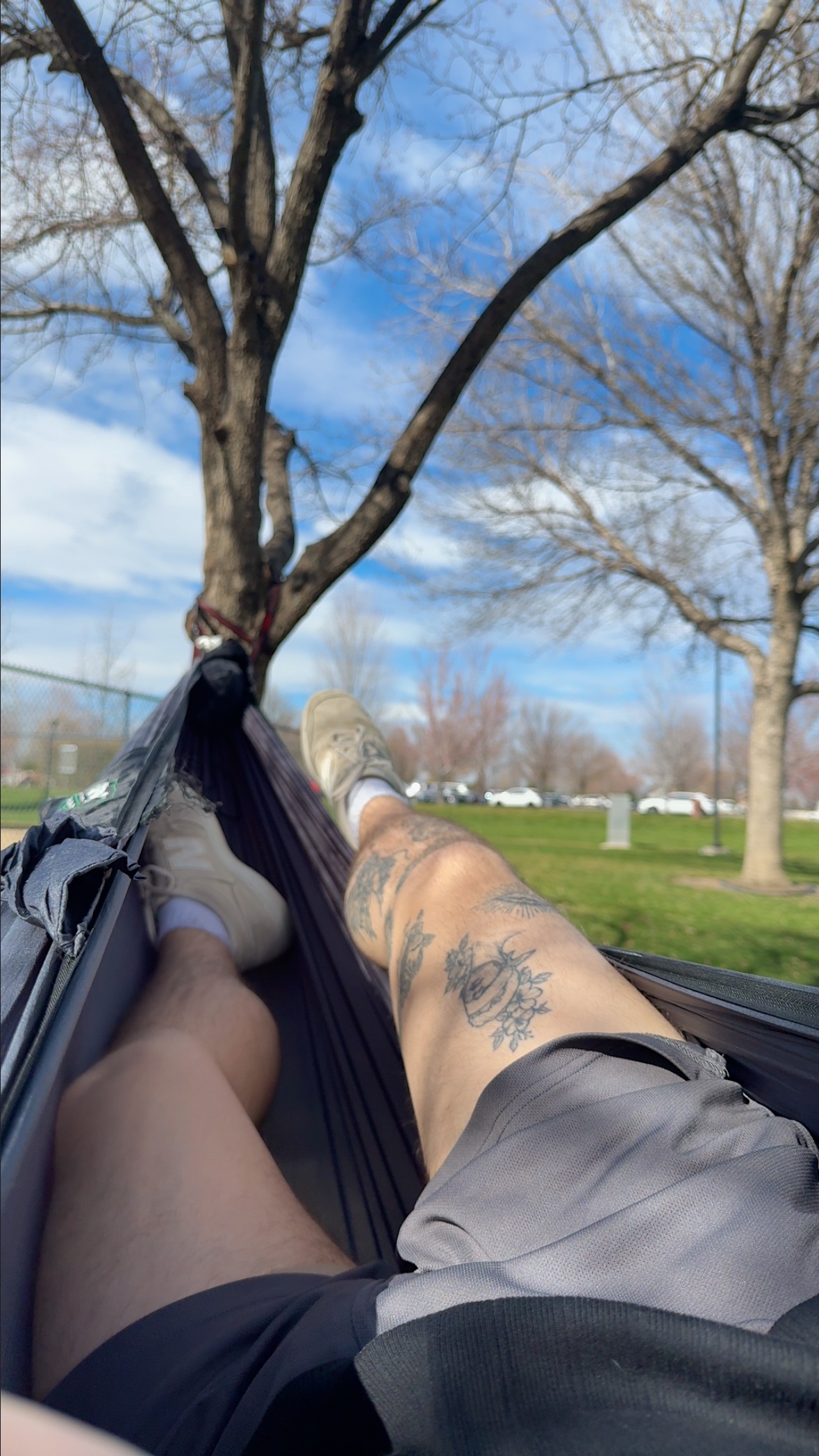 A person relaxes in a hammock in a park on a sunny day. Their legs are visible, showing off tattoos on one leg.