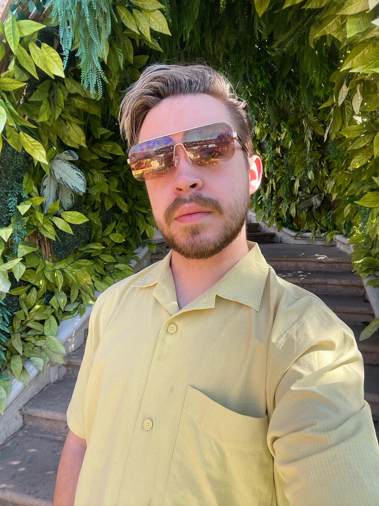 A man with sunglasses and stubble takes a selfie outdoors, wearing a light yellow button-up shirt. He is framed by lush green foliage and stairs behind him.