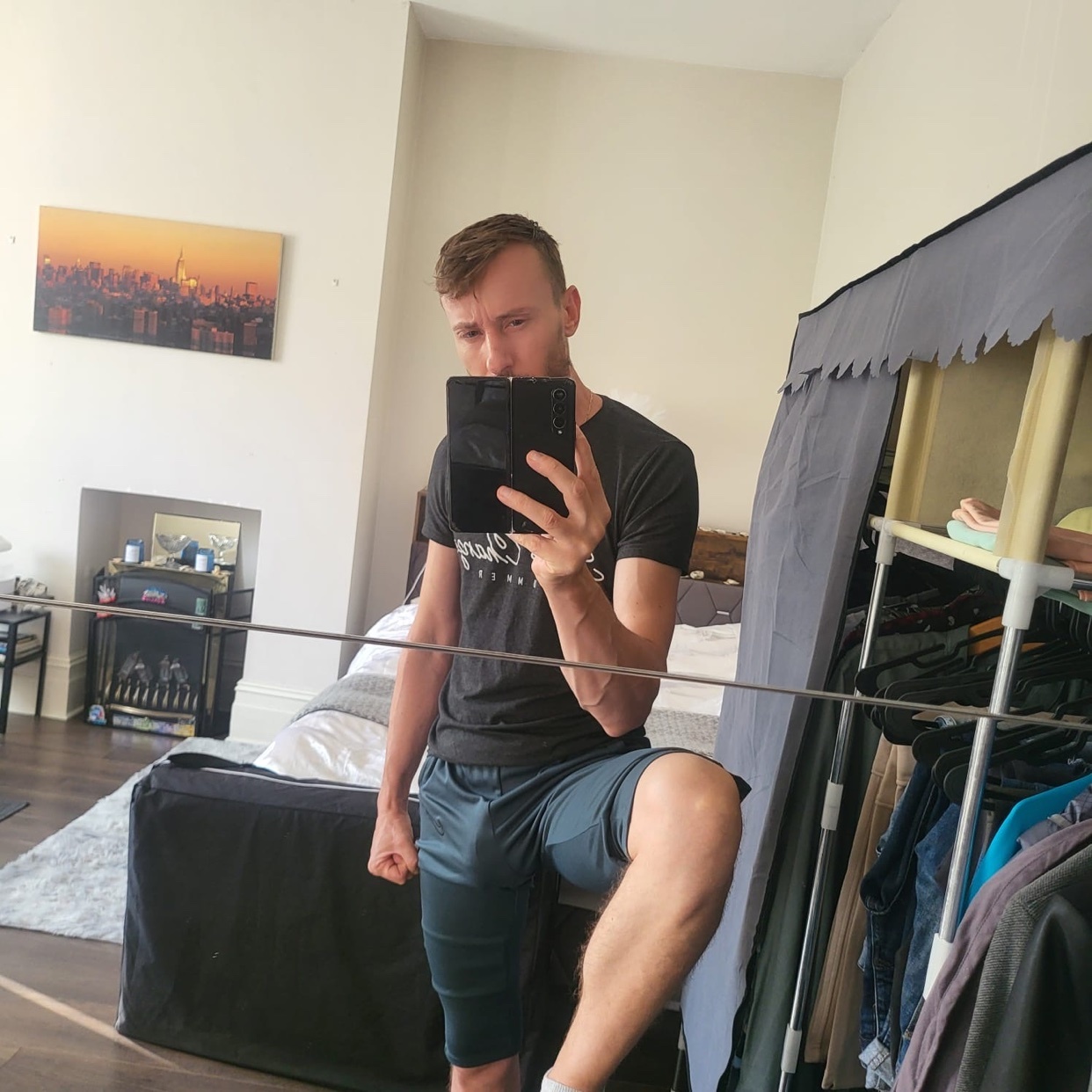 A man takes a mirror selfie in a bedroom, wearing a t-shirt and athletic shorts.