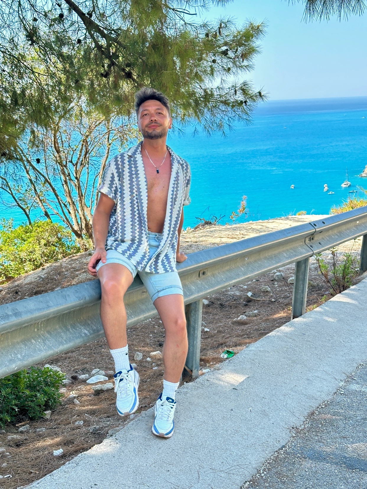A man poses on a guardrail with the ocean in the background. He is shirtless and wearing shorts and a patterned shirt.