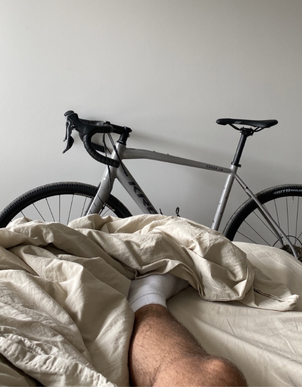 A bicycle is positioned behind a person's leg in a bed. The photo is taken from a low angle, emphasizing the minimalist aesthetic of the room.