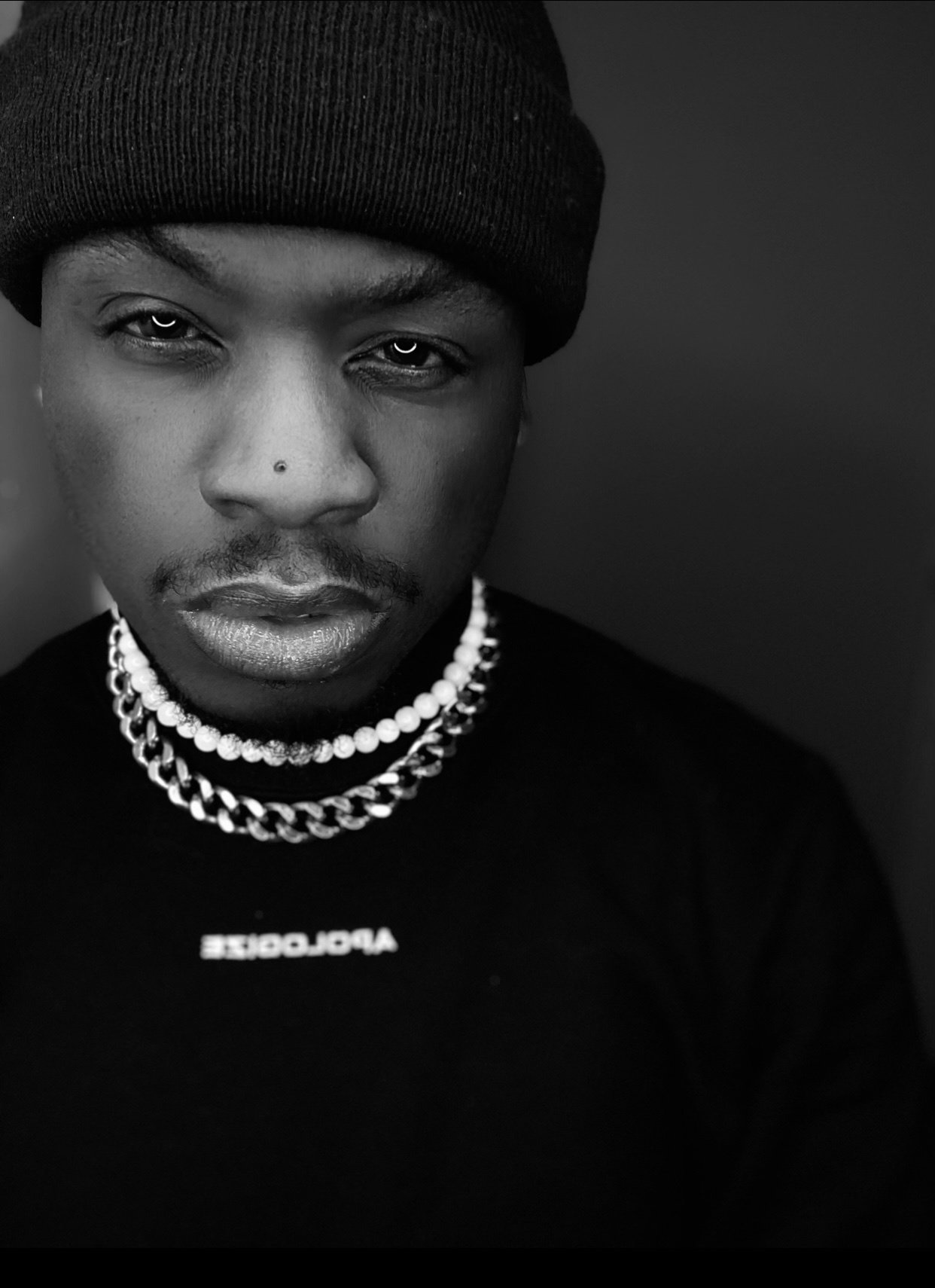 A black and white portrait of a man wearing a beanie and a necklace. He is looking directly at the camera.