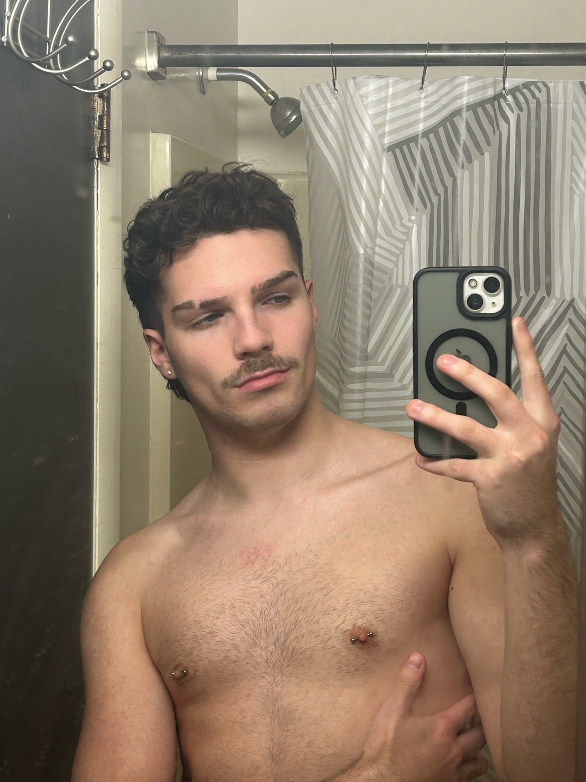 A shirtless man takes a mirror selfie in a bathroom. He has dark curly hair and a mustache.
