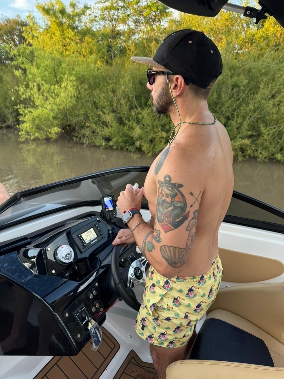 A man steers a boat on a sunny day. He is shirtless, showing off his tattoos and muscular physique.