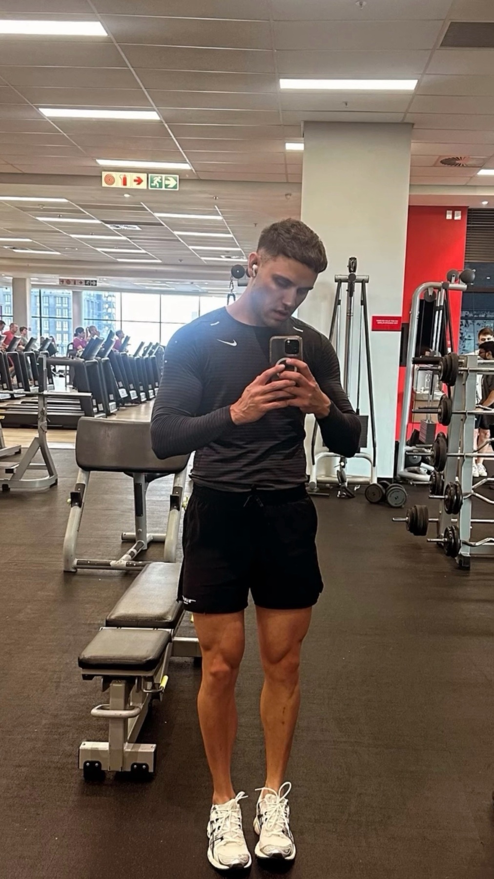 A man takes a mirror selfie at the gym. He is wearing a black long-sleeved shirt and shorts.