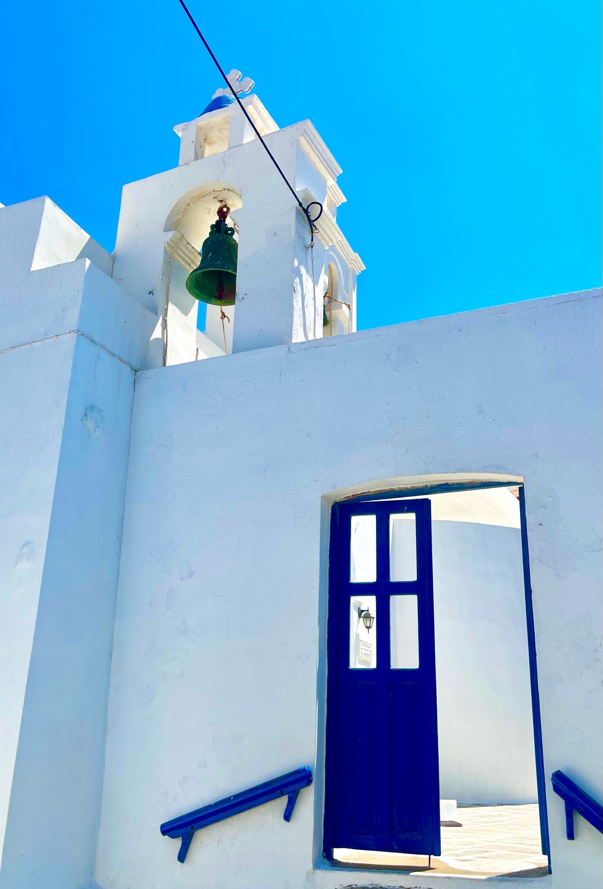 A bright, sunny day highlights a white Greek Orthodox church with a vibrant blue door and a bell tower against a clear blue sky. The bell hangs from a wire, ready to ring.