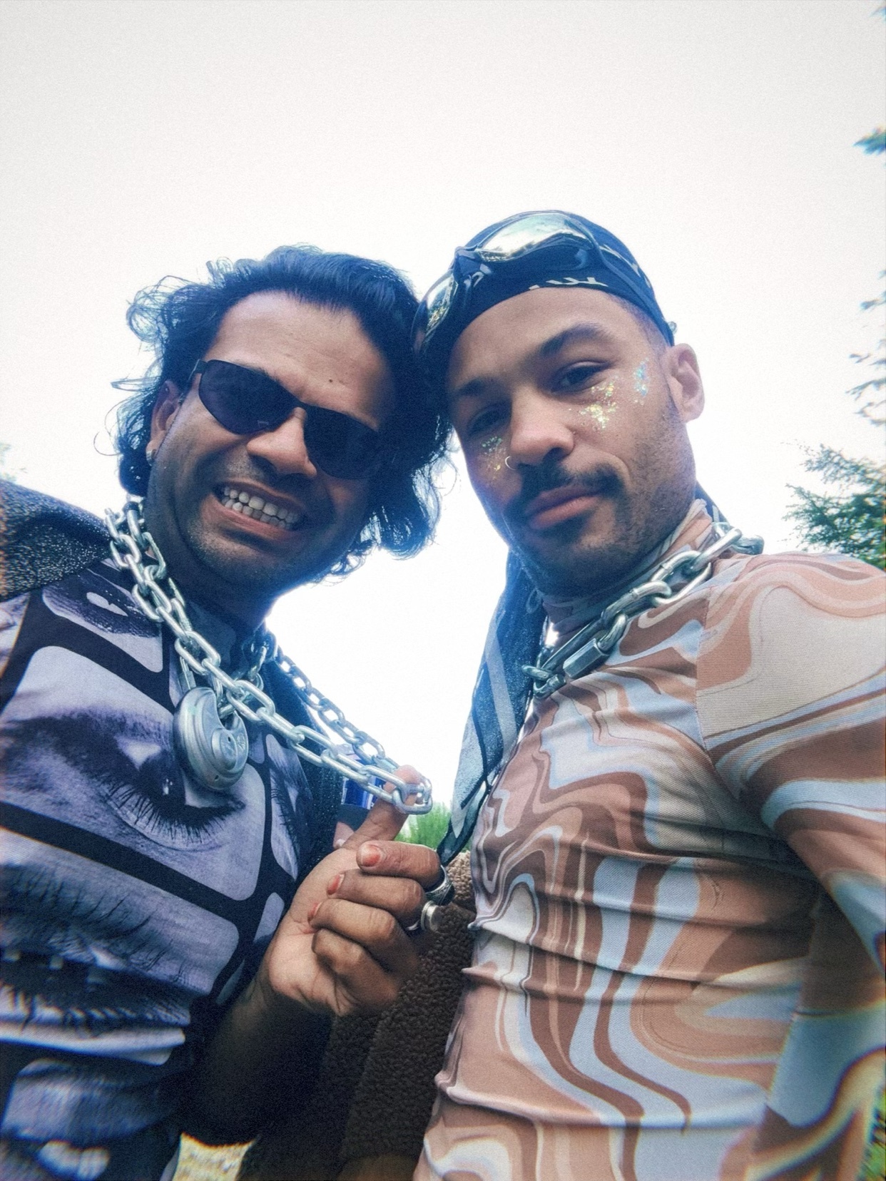 Two men pose for a selfie outdoors, both wearing unique patterned shirts and large chain necklaces. One man wears sunglasses and has glitter on his face.